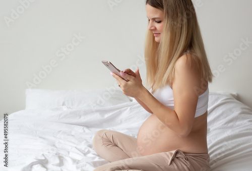 Portrait. A pregnant blonde sits on a bed and calls on the phone. Work at home. Close-up. High quality photo. Copyspace 