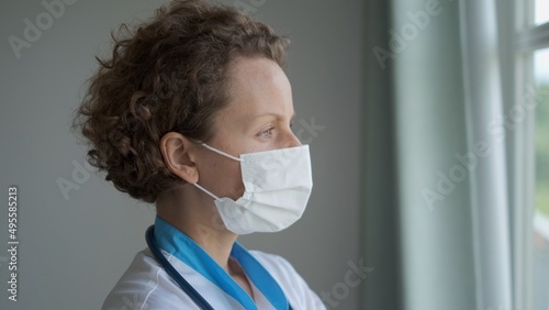 A woman doctor in a mask stands at the window, thinking about the severity of the disease of the patient whom she treats. Treatment of patients infected with a pulmonary infection. Tired nurse.
