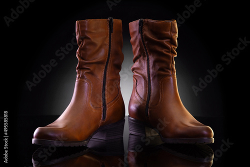 Pair of unbranded brown leather women’s mid-calf boots on black reflective surface. Product photography and shopping concept