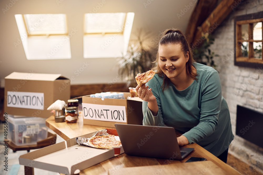 Charity community worker works on laptop and eats pizza while organizing donation boxes. Charity community worker works on laptop and eats pizza while organizing donation boxes.