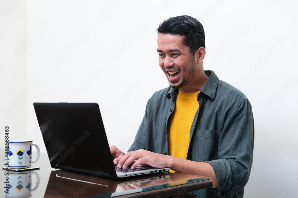 Adult Asian man showing excited expression when typing on his laptop Adult Asian man showing excited expression when typing on his laptop