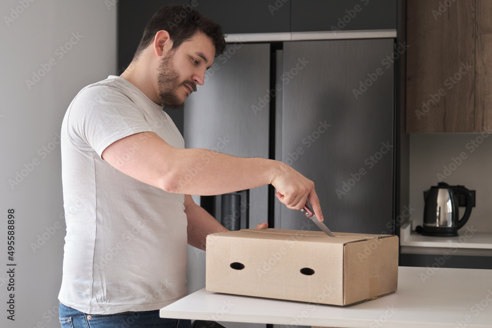Young latin man opening a cardboard box using a knife in the kitchen. Young latin man opening a cardboard box using a knife in the kitchen.