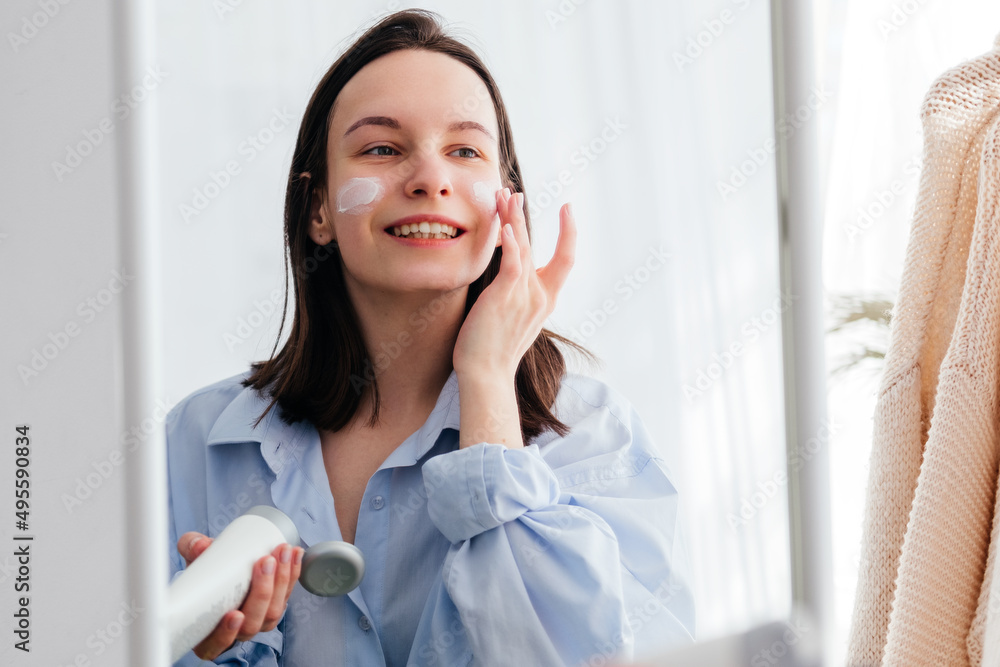 Caucasian young woman touching face and applying cream looking in mirror indoors. Home beauty skincare concept Caucasian young woman touching face and applying cream looking in mirror indoors. Home beauty skincare concept
