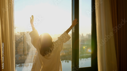 Young woman in white bathrobe open curtains in bedroom after waking up. Lady stretching with rising hands, looking out panoramic window enjoying sunrise in early morning