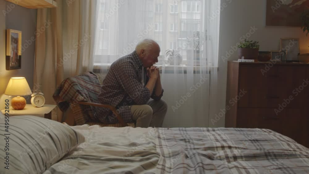 Medium long of sad aged Caucasian man with mustache sitting in armchair in his bedroom early in morning, looking out window and thinking Medium long of sad aged Caucasian man with mustache sitting in armchair in his bedroom early in morning, looking out window and thinking