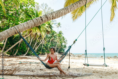 Young woman wearing red bikini relaxing in the hammock on sandy beach with coconut palm trees