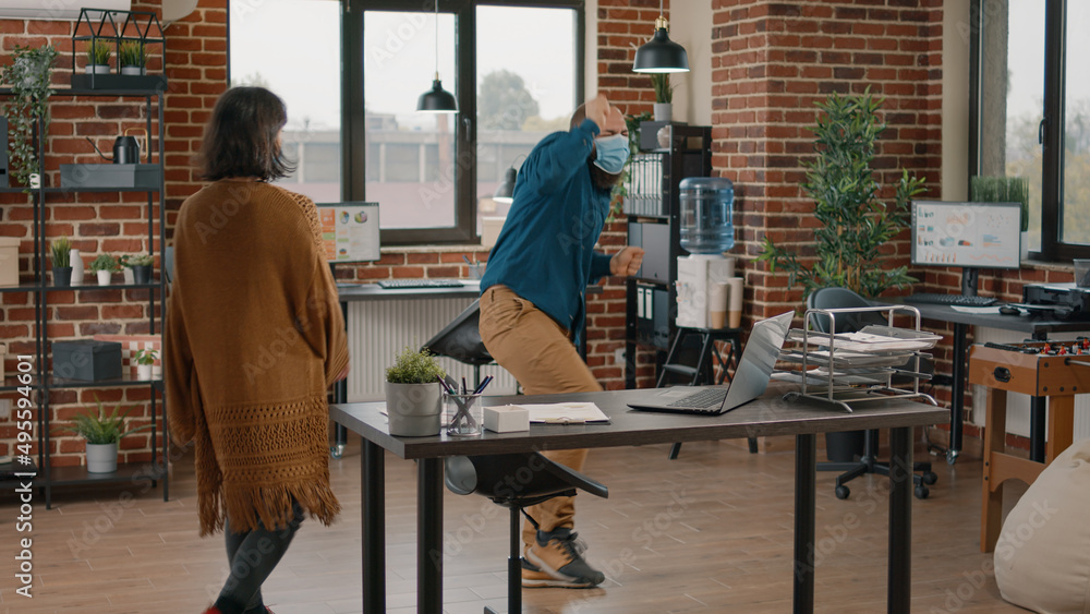 Man with face mask doing funny dance moves and celebrating victory after reading good news on laptop. Woman catching colleagues dancing and celebrating achievement in startup office. Man with face mask doing funny dance moves and celebrating victory after reading good news on laptop. Woman catching colleagues dancing and celebrating achievement in startup office.