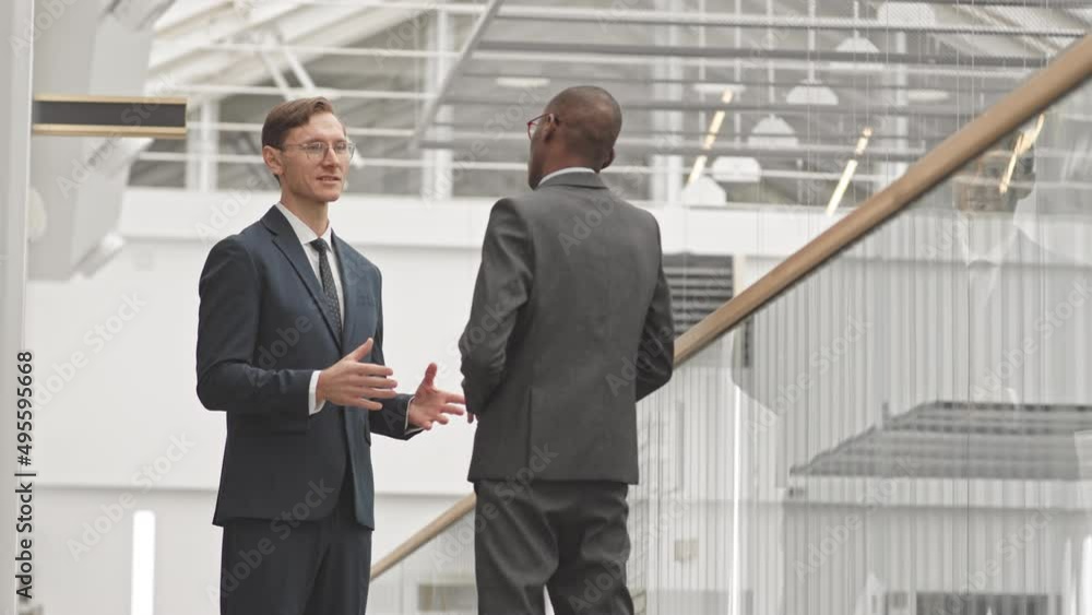Medium slowmo of two young successful multiethnic businessmen shaking hands after making deal, standing by glass railing at modern business center Medium slowmo of two young successful multiethnic businessmen shaking hands after making deal, standing by glass railing at modern business center