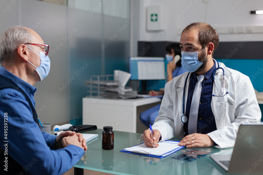 Doctor consulting mature man and taking notes at checkup visit during pandemic. Specialist doing medical examination with patient, writing diagnosis on appointment form in cabinet. Doctor consulting mature man and taking notes at checkup visit during pandemic. Specialist doing medical examination with patient, writing diagnosis on appointment form in cabinet.