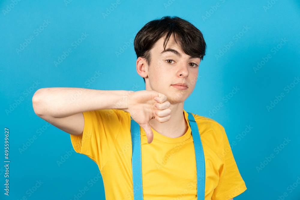 Cute teenager boy with thumbs down looking at camera isolated on blue background. Cute teenager boy with thumbs down looking at camera isolated on blue background.