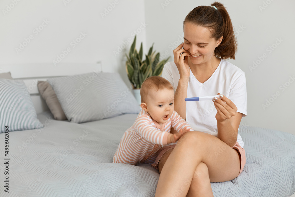 Indoor shot of smiling happy woman wearing white t shirt and shorts sitting on bed with her baby daughter and holding pregnancy test with positive result, calling to husband to say good news. Indoor shot of smiling happy woman wearing white t shirt and shorts sitting on bed with her baby daughter and holding pregnancy test with positive result, calling to husband to say good news.