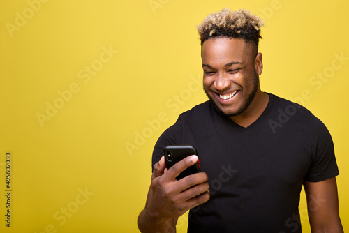 A curly joyful man reads the news on his phone. A dark-skinned strong young man smiles looking at the screen of a smartphone, on a yellow background in a black T-shirt © Aleksandr