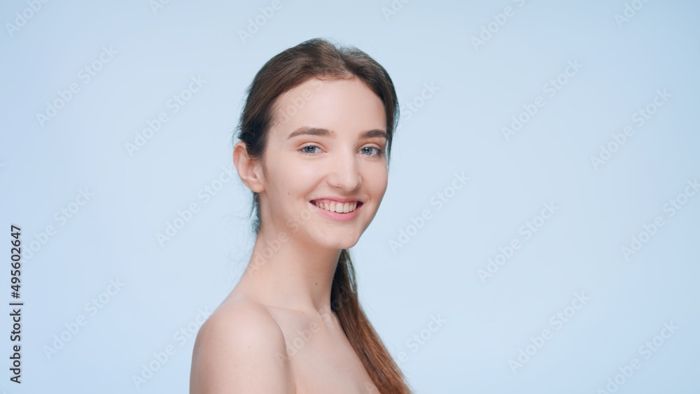 Young attractive brunette woman with a ponytail looks at camera over her shoulder and smiles widely on blue background | Skin care and beauty care product commercial concept Young attractive brunette woman with a ponytail looks at camera over her shoulder and smiles widely on blue background | Skin care and beauty care product commercial concept