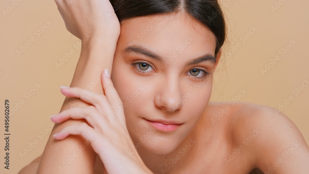 Close-up beauty portrait of young woman looks at camera and softly touches her hand skin, raises up her chin and smiles | Skincare products promotional concept