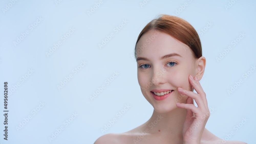 Close-up beauty portrait with place for sign or product. Young ginger woman gently touches her face sliding hand from the chin and gently lays fingers at cheek and jawline, looks at camera and smiles Close-up beauty portrait with place for sign or product. Young ginger woman gently touches her face sliding hand from the chin and gently lays fingers at cheek and jawline, looks at camera and smiles