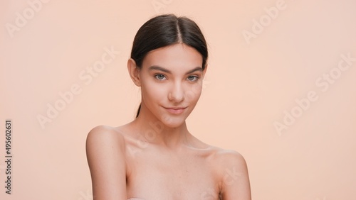 Medium close-up beauty portrait of young slim woman looking at the camera lifting up her shoulder on nude background | Beauty care concept.