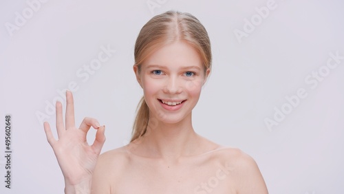 Young blonde woman makes an ok sign, looks at the camera and smiles.