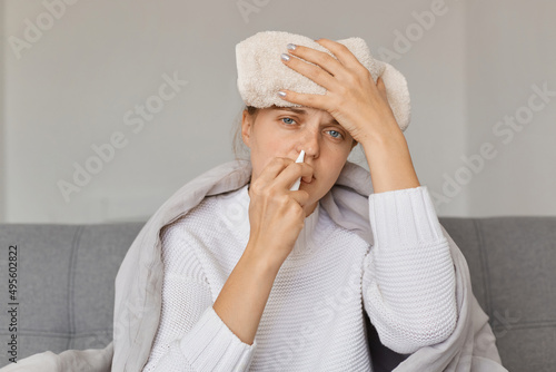 Portrait of sick upset woman with wet towel on her forehead being wrapped in blanket sitting on sofa and using nasal spray, treating runny nose, having high temperature.