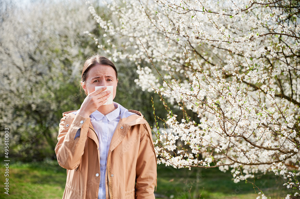 Woman allergic suffering from seasonal allergy at spring, posing in blossoming garden at springtime.Young woman sneezing and blowing nose in front of blooming tree. Spring allergy concept Woman allergic suffering from seasonal allergy at spring, posing in blossoming garden at springtime.Young woman sneezing and blowing nose in front of blooming tree. Spring allergy concept