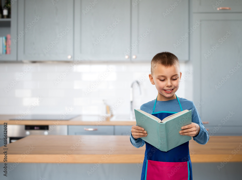 cooking, culinary and profession concept - happy smiling little boy in apron reading cook book over kitchen background cooking, culinary and profession concept - happy smiling little boy in apron reading cook book over kitchen background