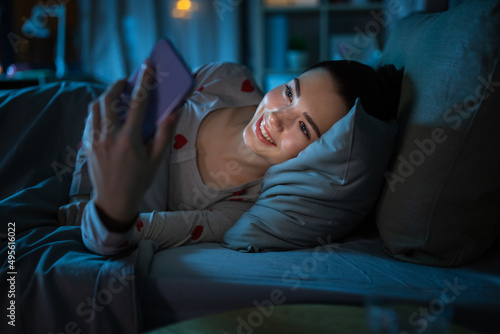 technology, bedtime and rest concept - smiling teenage girl in pajamas with smartphone lying in bed at night