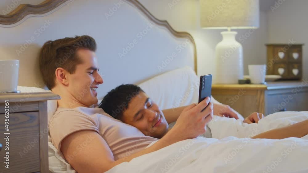 Loving same sex male couple lying in bed at home with man looking at social media on mobile phone whilst partner sleeps - shot in slow motion Loving same sex male couple lying in bed at home with man looking at social media on mobile phone whilst partner sleeps - shot in slow motion
