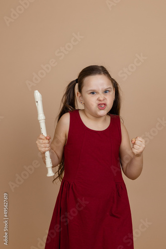 Girl in red dress with angry facial expression hold flute tightly in fist, beige background. Learning to play woodwind musical instrument. Flute and children is concept of music education development. photo