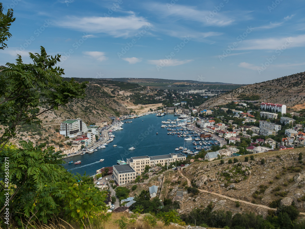 View from the mountain to the city with a pier and a bay. City in the mountains. Around the green foliage, trees, blue sky. Yachts, boats, buildings, houses are visible in the city. Urban landscape.
