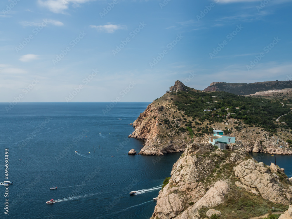 Mountain landscape. View of the neighboring mountains and the sea. There is a house at the top of the mountain. Boats, yachts go on the sea. They leave behind a trail of waves.