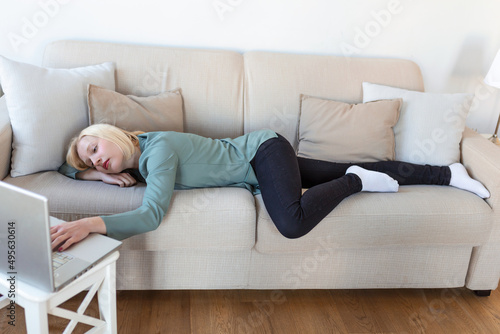Exhausted young woman lying on sofa, using laptop, too tired or bored of online work at home, free space. Workaholism, chronic fatigue, overworking on remote job concept