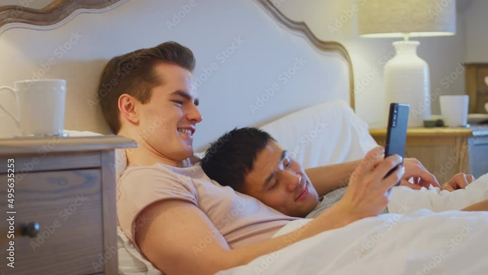 Loving same sex male couple lying in bed at home with man taking selfie on mobile phone whilst partner sleeps - shot in slow motion Loving same sex male couple lying in bed at home with man taking selfie on mobile phone whilst partner sleeps - shot in slow motion