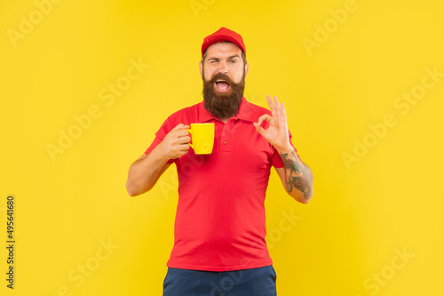 Happy winking guy in casual red cap and tshirt giving OK holding cup yellow background, coffee