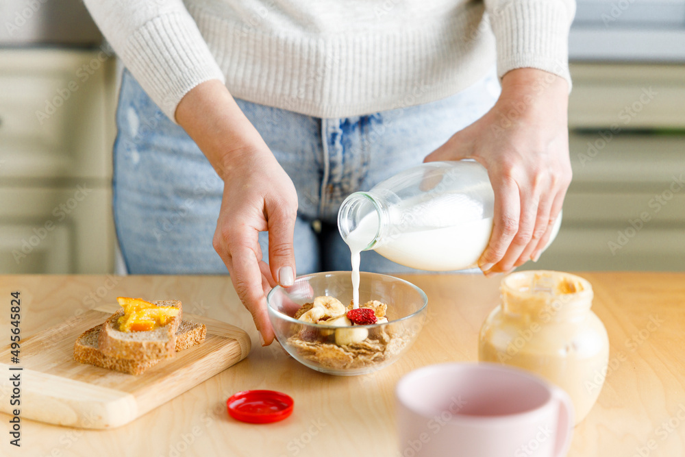 Young woman preparing healthy breakfast, pouring milk over cereals on the kitchen counter