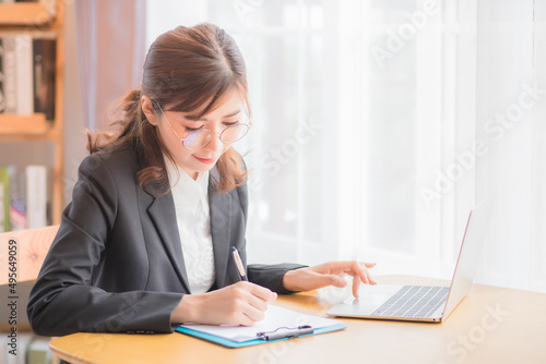 A Thai Southeast Asian businesswoman wearing a black suit is sitting on the desk typing notebooks and taking notes on her office desk.