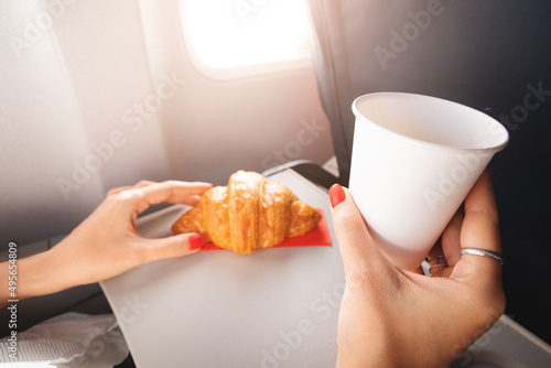 A girl passenger eats a croissant and drinks coffee sitting by the window on an airplane and flies on vacation photo