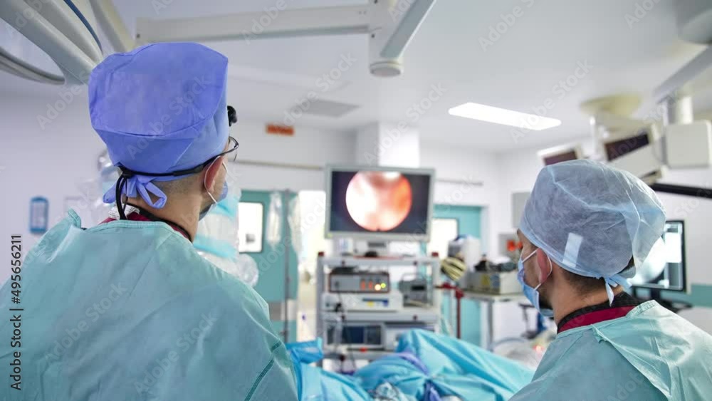 Surgeon standing their backs to the camera and looking at the screen. Monitor showing operational process in blur at backdrop. Surgeon standing their backs to the camera and looking at the screen. Monitor showing operational process in blur at backdrop.
