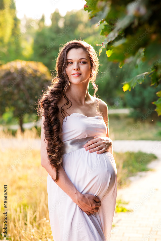 Young beautiful happy joyful pregnant woman in light pink long evening dress posing. background of blooming trees. Garden green leaves white flowers. Girl model. Elegance pregnancy spring fresh nature Young beautiful happy joyful pregnant woman in light pink long evening dress posing. background of blooming trees. Garden green leaves white flowers. Girl model. Elegance pregnancy spring fresh nature