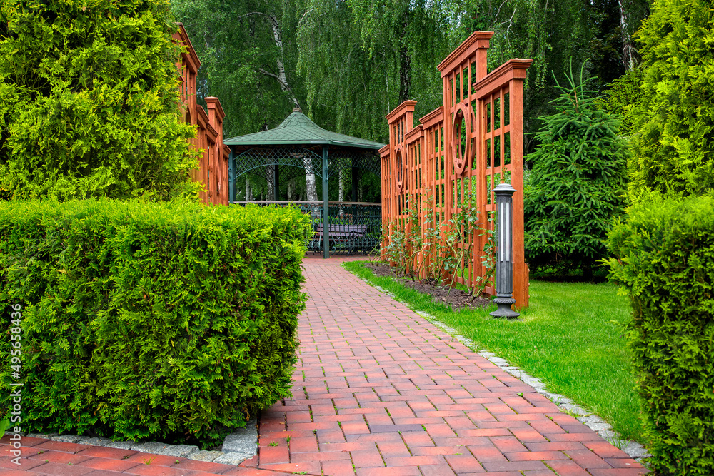 Vertical pergola made of brown wood in a roses garden with a stone tile walkway with granite curb and ground lantern in backyard in a garden with plants and thuja hedge with iron gazebo.