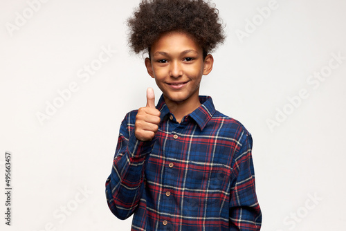 Body language, signs, gestures. Handsome teen boy with dark skin standing in front of camera with cute adorable sweet smile putting right thumb up, showing his excellent mood, good attitude