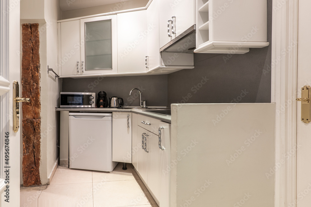 corner of a kitchen with white wooden cabinets, a glass cabinet, a gray countertop, a small white fridge and a microwave coffee maker and oven, a kettle on the counter corner of a kitchen with white wooden cabinets, a glass cabinet, a gray countertop, a small white fridge and a microwave coffee maker and oven, a kettle on the counter