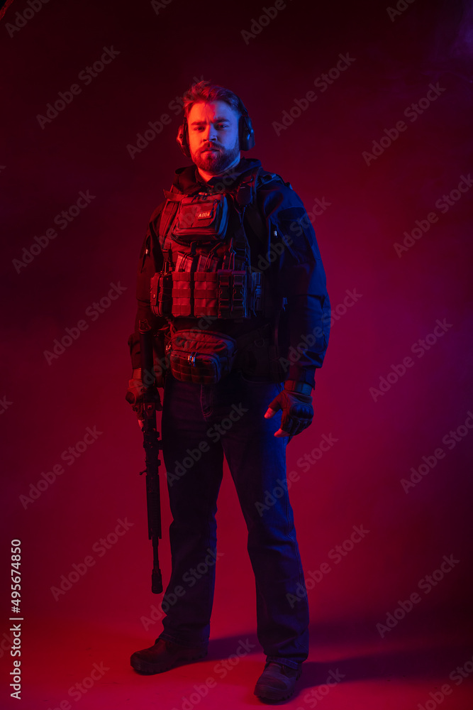 Fototapeta premium a fighter in equipment, body armor, headphones holds a machine gun, aims at the target, hits the target through the sight. military business concept. red background. blue-red light