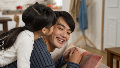 cute asian girl hugging and kissing her father’s face from behind on the sofa while he is lying relaxing. she sits and plays on the smiling man’s back in the living room at home © PRPicturesProduction