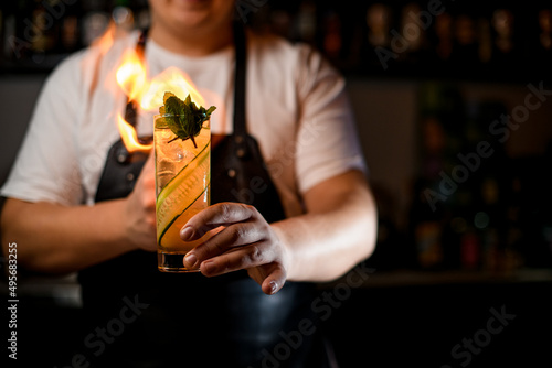 male hand holds glass of cocktail with cucumber slices and mint and sprinkles and sets it on fire
