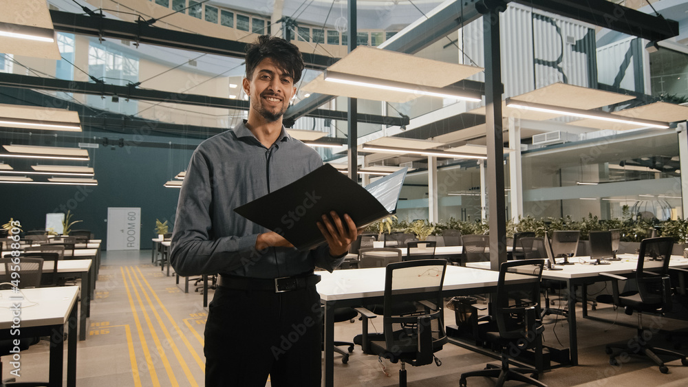 Young indian latin manager male business man employee checking document folder inspecting financial report raising head with satisfied look winking eye approvingly giving consent working agreement Young indian latin manager male business man employee checking document folder inspecting financial report raising head with satisfied look winking eye approvingly giving consent working agreement