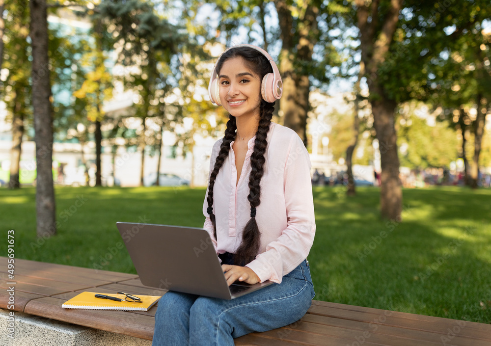 Happy indian lady relaxing with laptop and headphones in park, sitting on bench and smiling at camera outdoors Happy indian lady relaxing with laptop and headphones in park, sitting on bench and smiling at camera outdoors