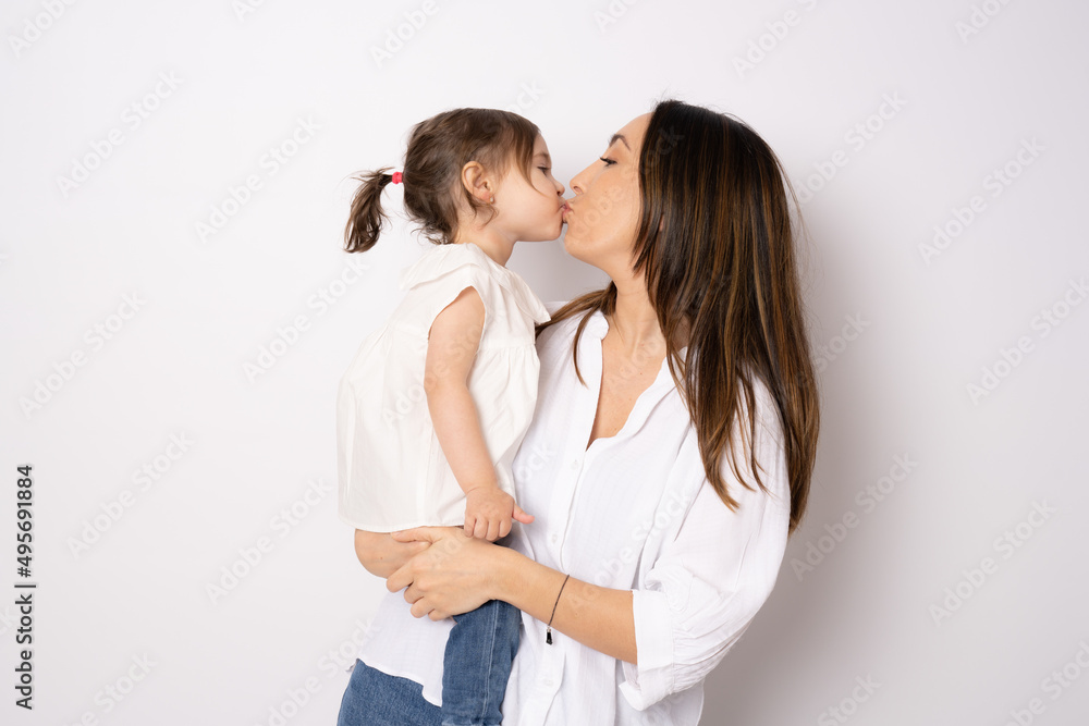 Photo of pretty young mommy kissing little daughter standing isolated over white background. Motherhood concept. Photo of pretty young mommy kissing little daughter standing isolated over white background. Motherhood concept.