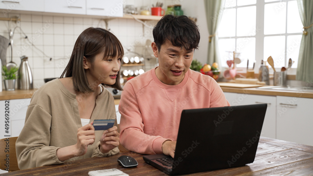 happy asian married couple enjoying shopping online with credit card and laptop. the man is keying in the number to make payment as the woman is reading to him happy asian married couple enjoying shopping online with credit card and laptop. the man is keying in the number to make payment as the woman is reading to him