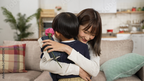 Asian baby boy show his mother the carnation flower and Mother’s Day gift. smiling mother giving her son a big hug in living room