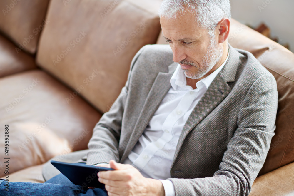 Reading an interesting article online. Shot of a handsome mature man using his tablet while sitting on the sofa at home. Reading an interesting article online. Shot of a handsome mature man using his tablet while sitting on the sofa at home.