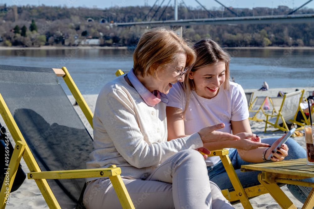 Mother and daughter spend quality time by the river together, bonding, drinking coffee, and looking at photos on their cell phone.